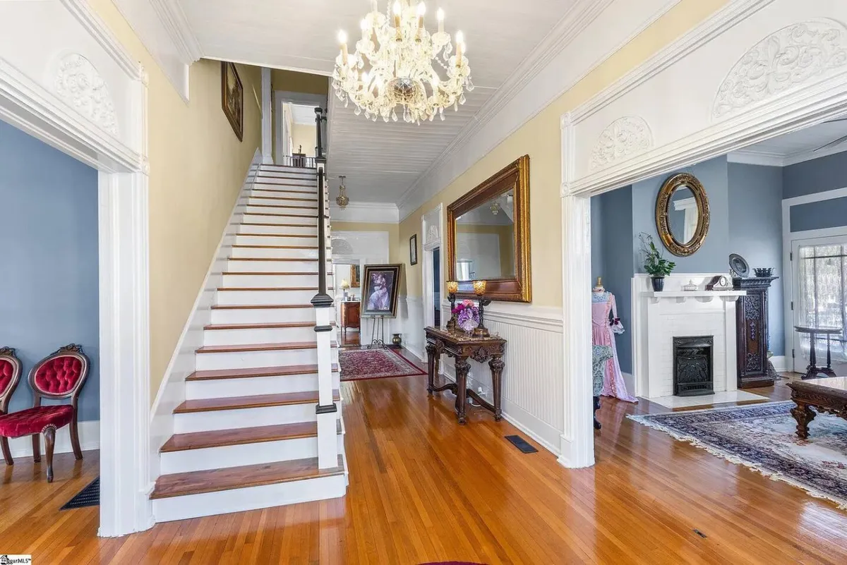 This image features a grand staircase with a striking chandelier above, a decorative mirror, and antique furniture. The scene exudes sophistication and warmth, capturing the spirit of a historic home where stories and gatherings unfold. The antique touches and curated décor elements are clearly visible, highlighting the unique character of the common areas.