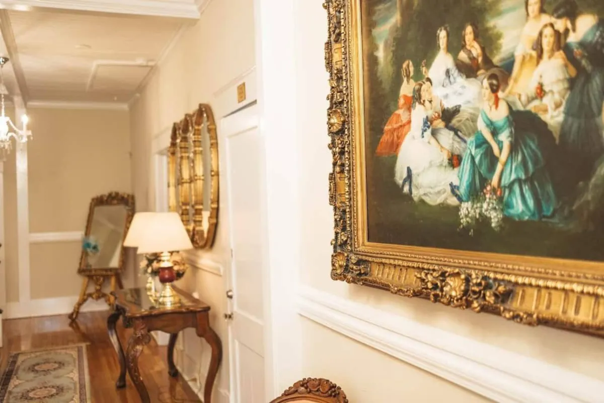 This image reveals an elegant interior space centered around an ornate painting, a sparkling chandelier, decorative mirrors, and an antique table, all set within what appears to be a formal dining area. Soft lighting and classic decor evoke an atmosphere of refined hospitality and sociable gatherings.