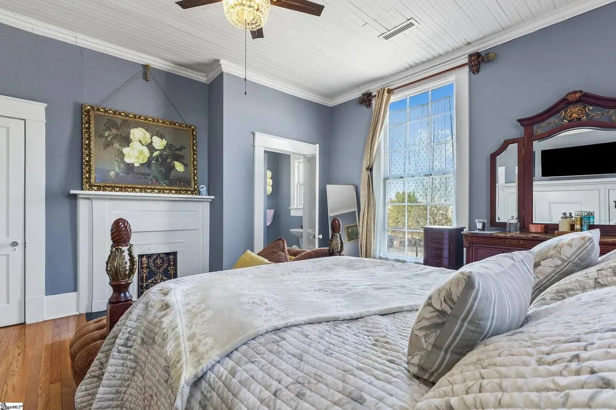 A beautifully decorated bedroom featuring a bed with plush bedding, an ornate mirror, and a decorative painting above the bed. Large windows allow abundant natural light to flood the room, enhancing its cozy yet elegant ambiance. The setting is perfect for enjoying a peaceful morning or winding down in the evening.