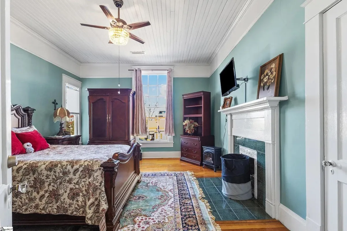 A serene bedroom scene with an ornate headboard, large window streaming in natural light, a fireplace with an elegant mantel, and a bookshelf displaying thoughtfully chosen décor. The overall ambiance is comfortable and inviting, with clear historic and personal touches evident throughout the space.