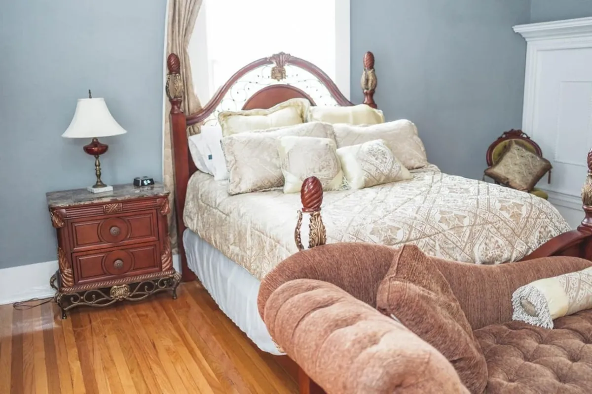 This image showcases an ornate bed dressed with soft, luxurious linens and decorative pillows. A vintage bedside table and a plush armchair sit nearby, bathed in gentle natural light that streams through the window. The atmosphere is cozy and elegant, making it feel like a tranquil retreat ideal for relaxing or reading.
