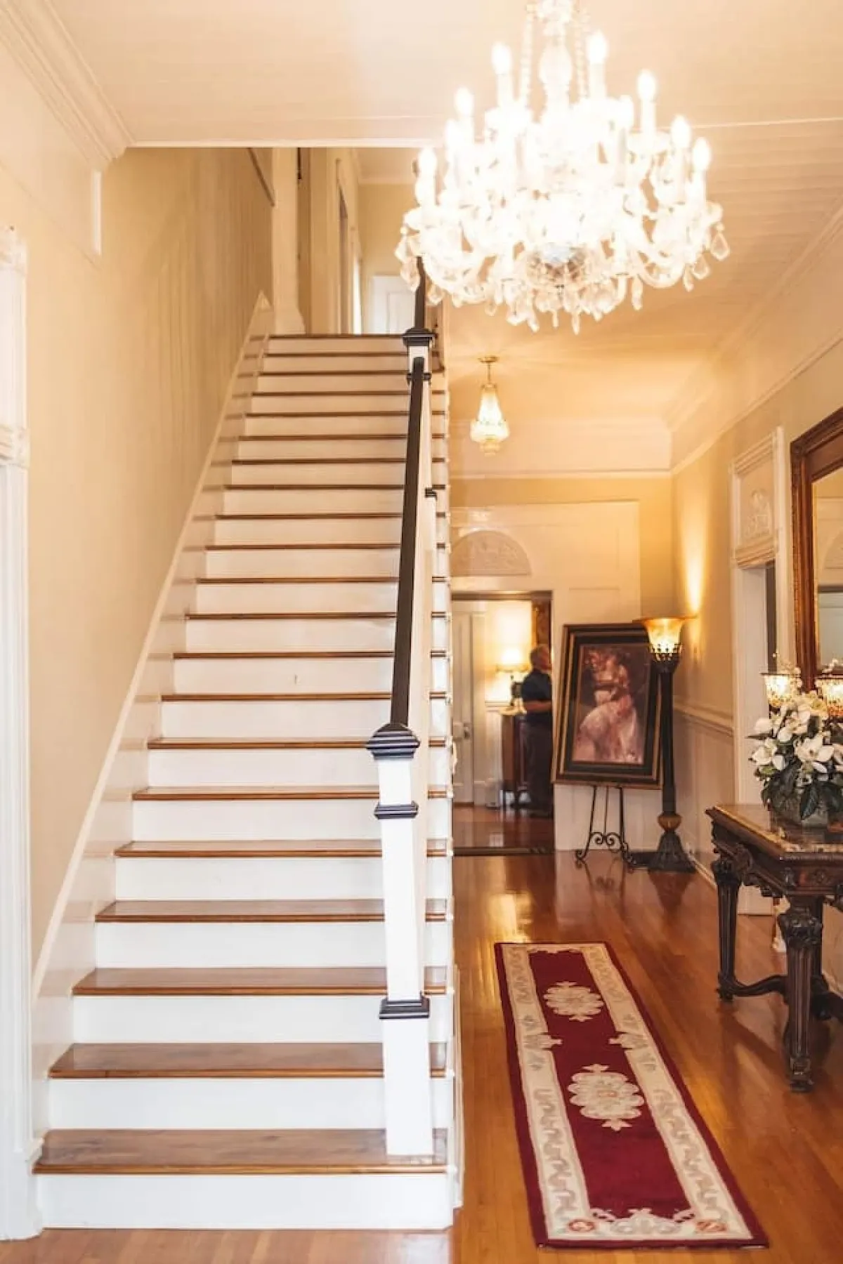 A graceful interior featuring a sweeping staircase, an ornate chandelier overhead, framed artwork, and a floral arrangement. The setting feels inviting and stately, with plenty of natural light and space for guests to gather or pass through on their way to breakfast.
