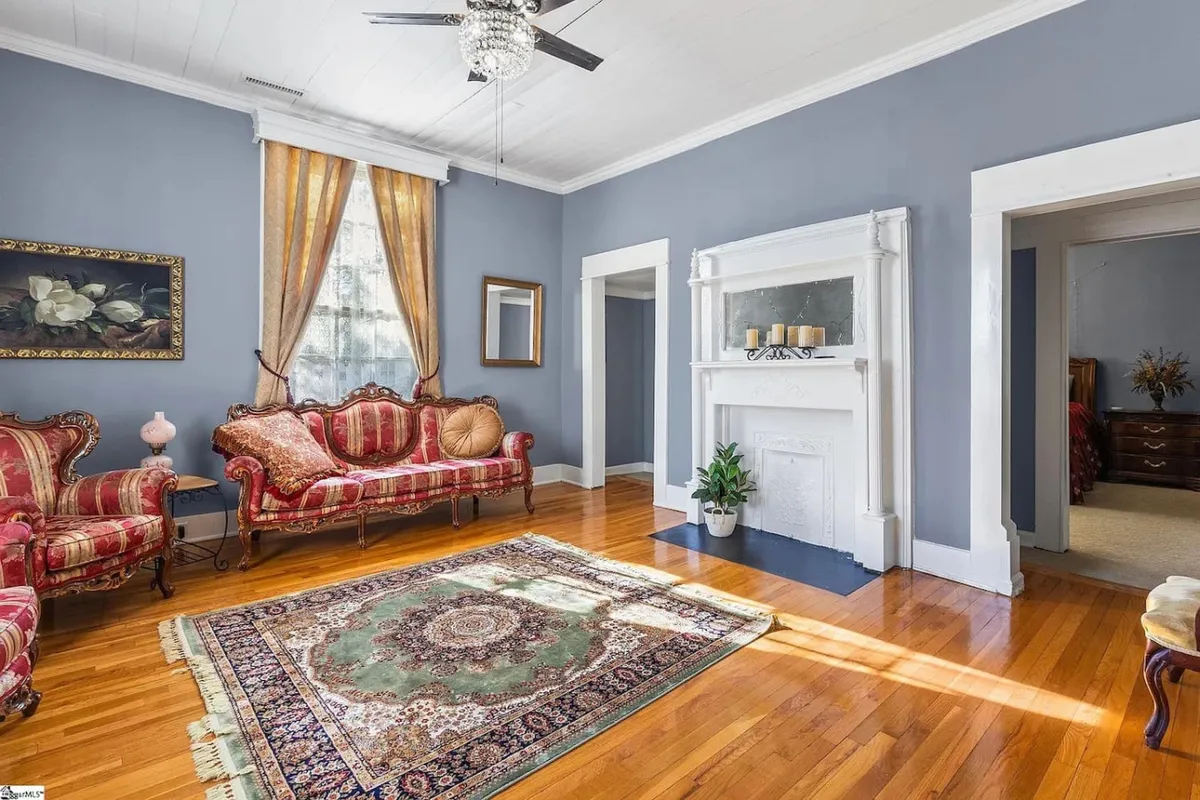 This image features a beautifully furnished living room with an ornate couch, a stately fireplace, and a decorative rug. The space emanates elegance and comfort, ideal for relaxing, reading, or intimate social moments. Soft, ambient lighting enhances the luxurious yet cozy feel of the room.