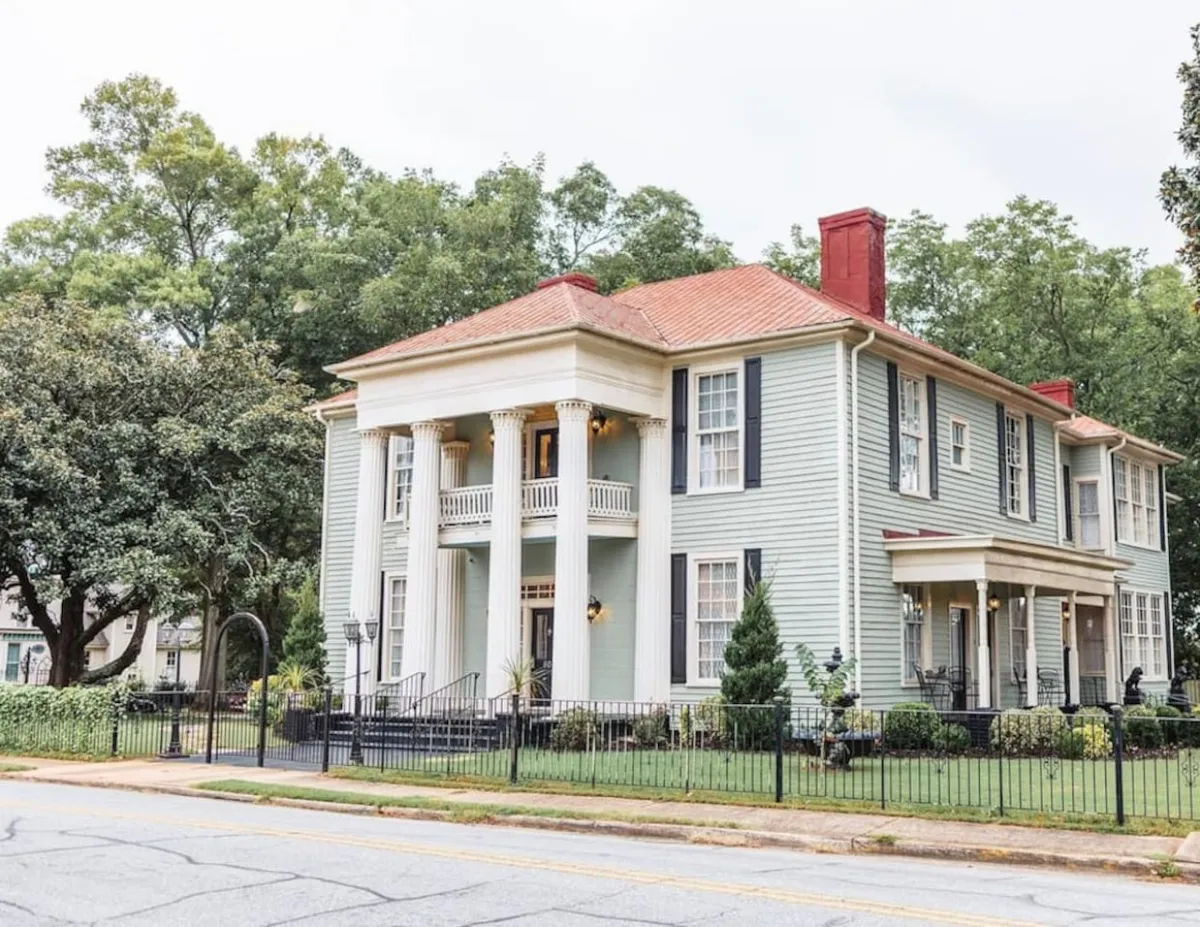 The image showcases the stately exterior of a historic single-family house with grand white columns, a striking red roof, a beautifully manicured lawn, and a charming decorative fence. The scene is bathed in warm sunset light, inviting guests to imagine relaxing on the porch or hosting elegant outdoor gatherings in the spacious yard.