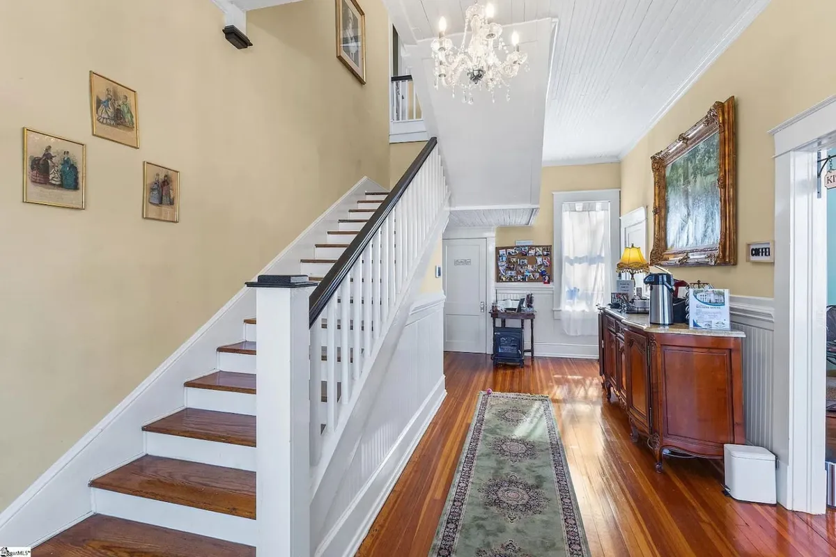 This image showcases an inviting interior space highlighted by a graceful staircase, a sparkling chandelier, a plush rug, elegant wall art, and comfortable furniture. The scene suggests the first steps into the home, warmth, and the ambiance of shared gatherings. The lighting is soft and natural, creating a sense of welcome and openness.