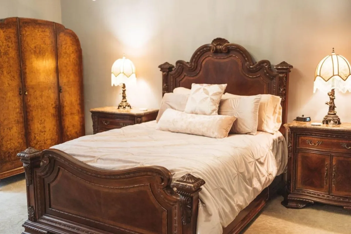 This inviting bedroom highlights an ornate bed frame, vintage lamps, and an antique wardrobe. Sunlight floods the space, creating an atmosphere that is both nostalgic and welcoming. The decor and furniture choices reflect curated taste and a nod to the area's rich heritage.